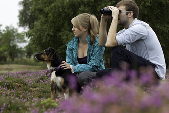 A young man and woman sit amongst flowering heather, with a dog on a lead sat beside them. The woman has her hands on the dog, whilst the man looks through a pair of binoculars.