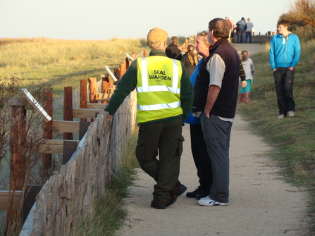 Seal Warden Volunteer stood against a fence with members of the public at Donna Nook